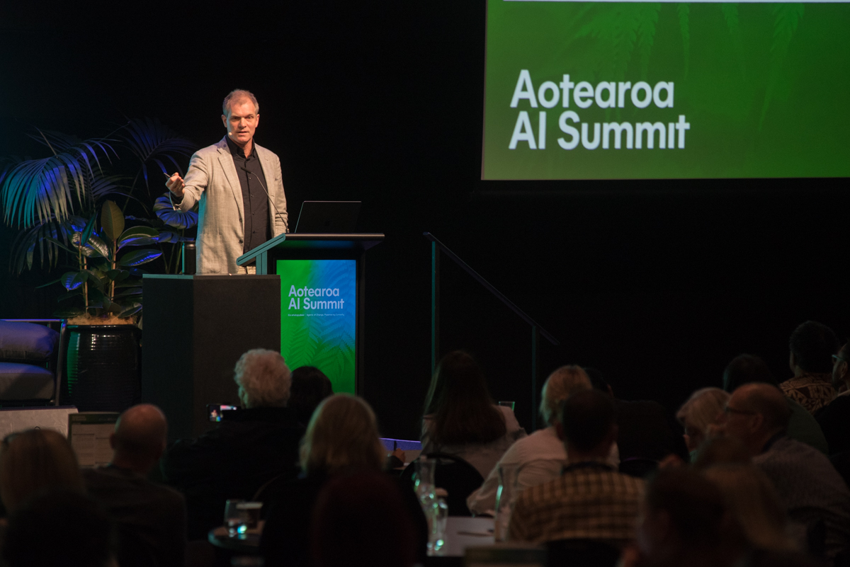 A speaker presenting from the podium at the Aotearoa AI Summit 2026, with the event logo on screen behind him and the audience seated in the foreground.