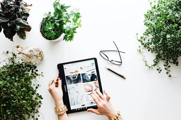 Top-down view of a white desk with potted plants, a tablet in use, glasses, pen, and coffee.