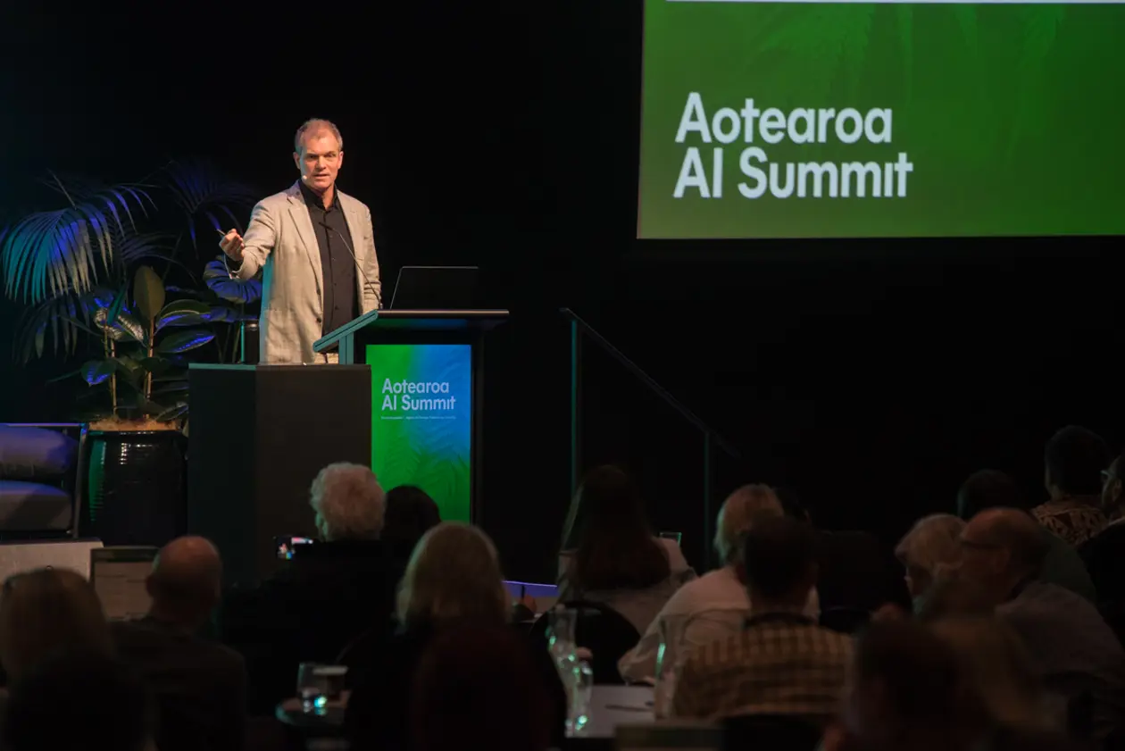 A speaker presenting from the podium at the Aotearoa AI Summit 2026, with the event logo on screen behind him and the audience seated in the foreground.