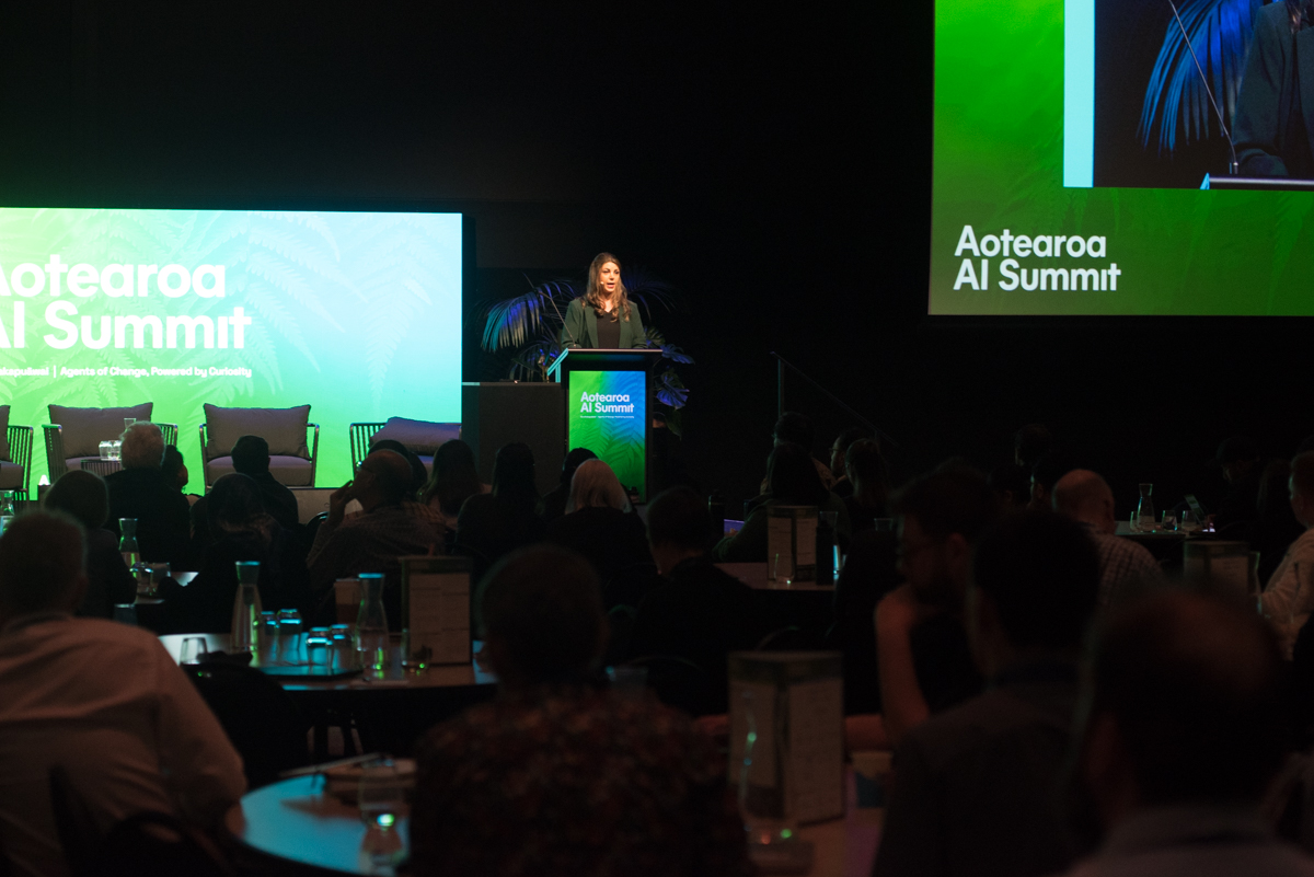 A speaker addresses the audience from a lectern at the 2026 Aotearoa AI Summit, with Aotearoa AI Summit branding on large green screens either side of the stage.