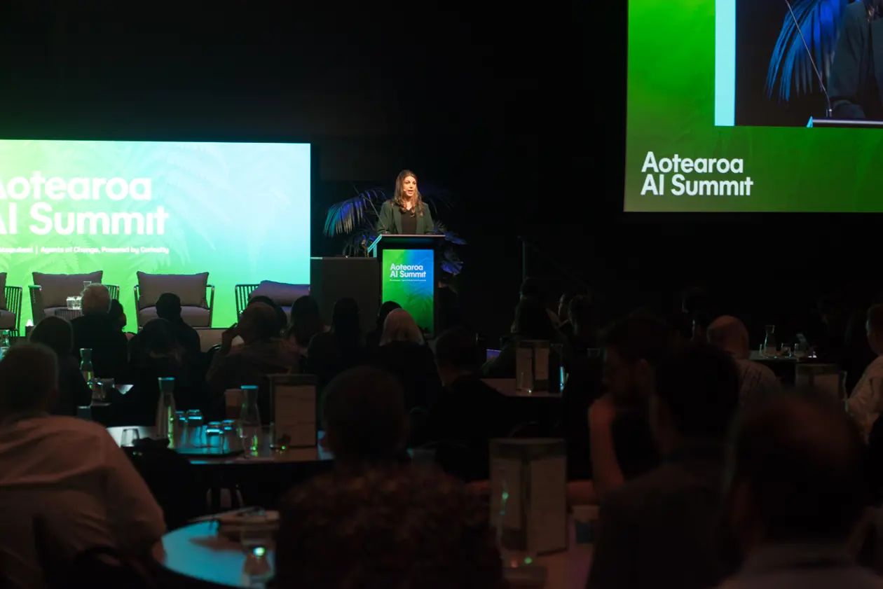 A speaker addresses the audience from a lectern at the 2026 Aotearoa AI Summit, with Aotearoa AI Summit branding on large green screens either side of the stage.