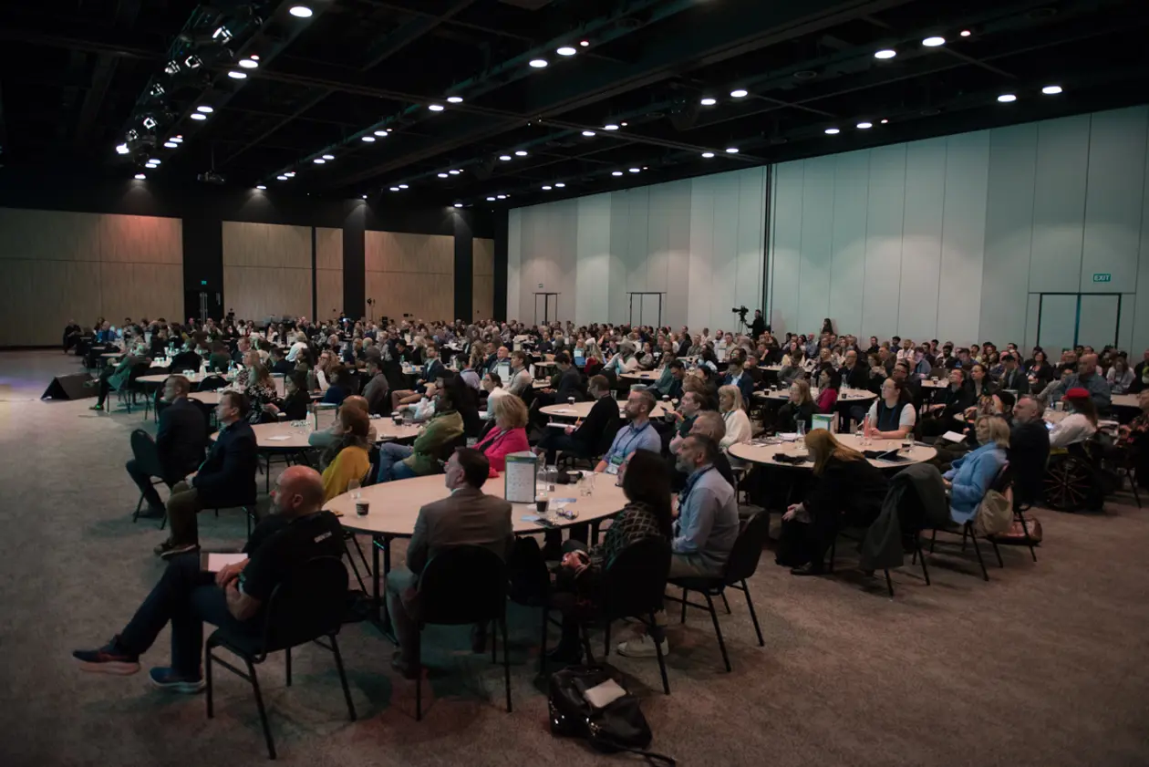 A packed conference hall at the Aotearoa AI Summit 2026, with attendees seated at round tables watching a session.